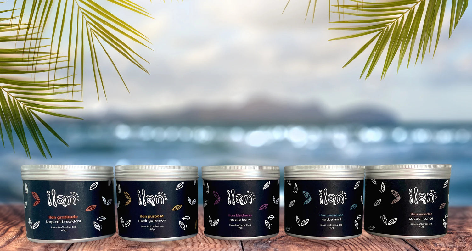 Five round metal tins of Nari body butter with silver lids and dark navy labels are lined up on a wooden surface with a blurred beach background and palm leaves framing the top of the image.